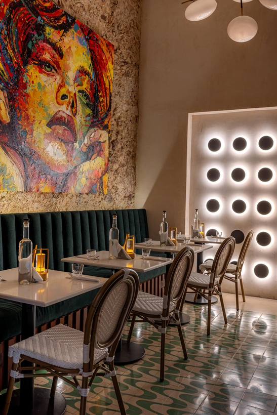 Comedor con banca de terciopelo verde, sillas de bistró, un gran mural y un muro de luces circulares